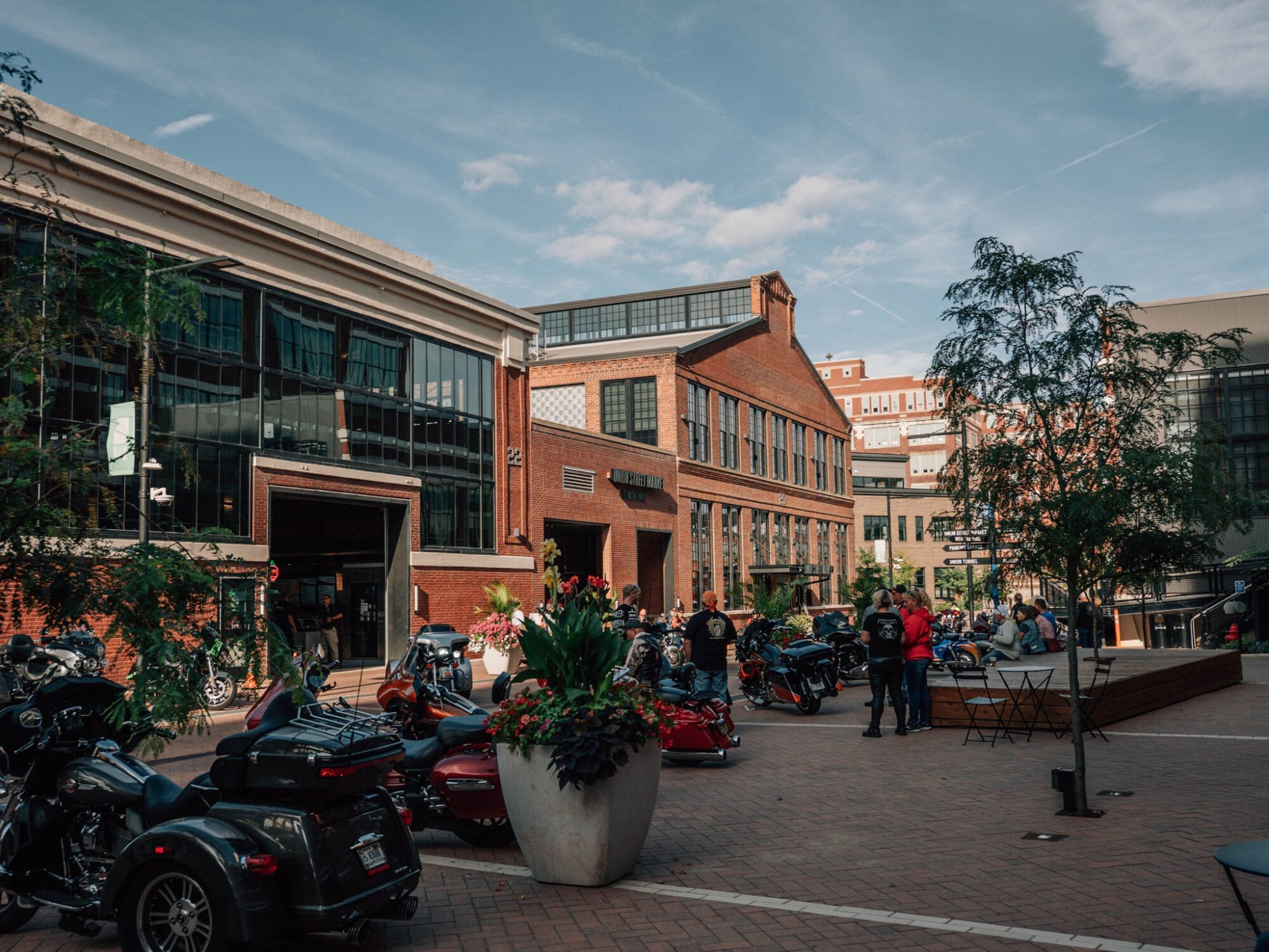 Urban plaza with brick buildings, many parked motorcycles, and a few people near a large flower-filled planter under a blue sky.