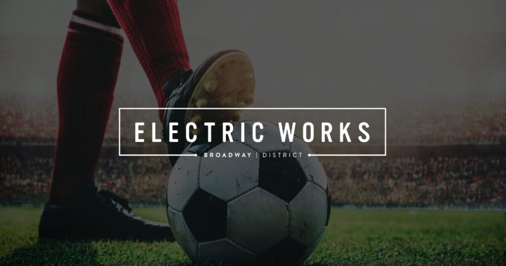 Soccer ball on a grass field with a player's foot about to kick; white 'Electric Works' logo centered over a stadium backdrop.